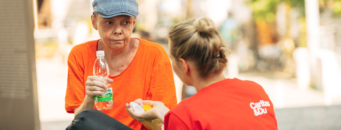 Eine Caritas-Mitarbeiterin reicht einer Frau, die eine Wasserflasche in der Hand hält, ein Hitzepaket.